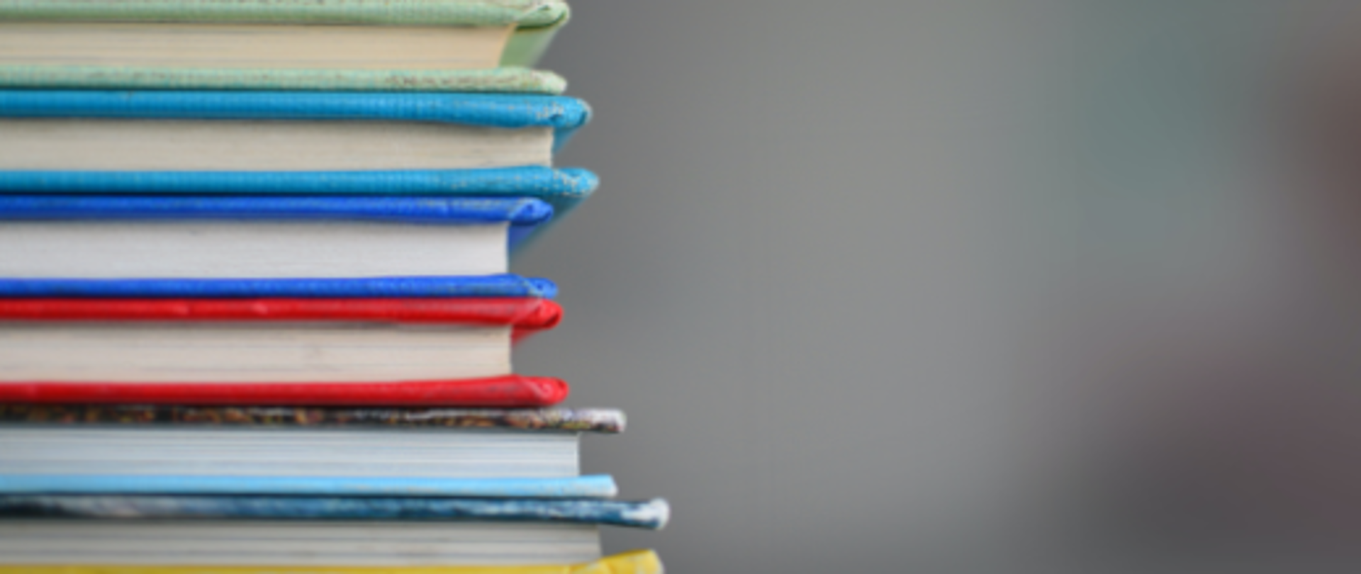 Fotografia przedstawia stos kolorowych książek na rozmytym, nieostrym szarym tle