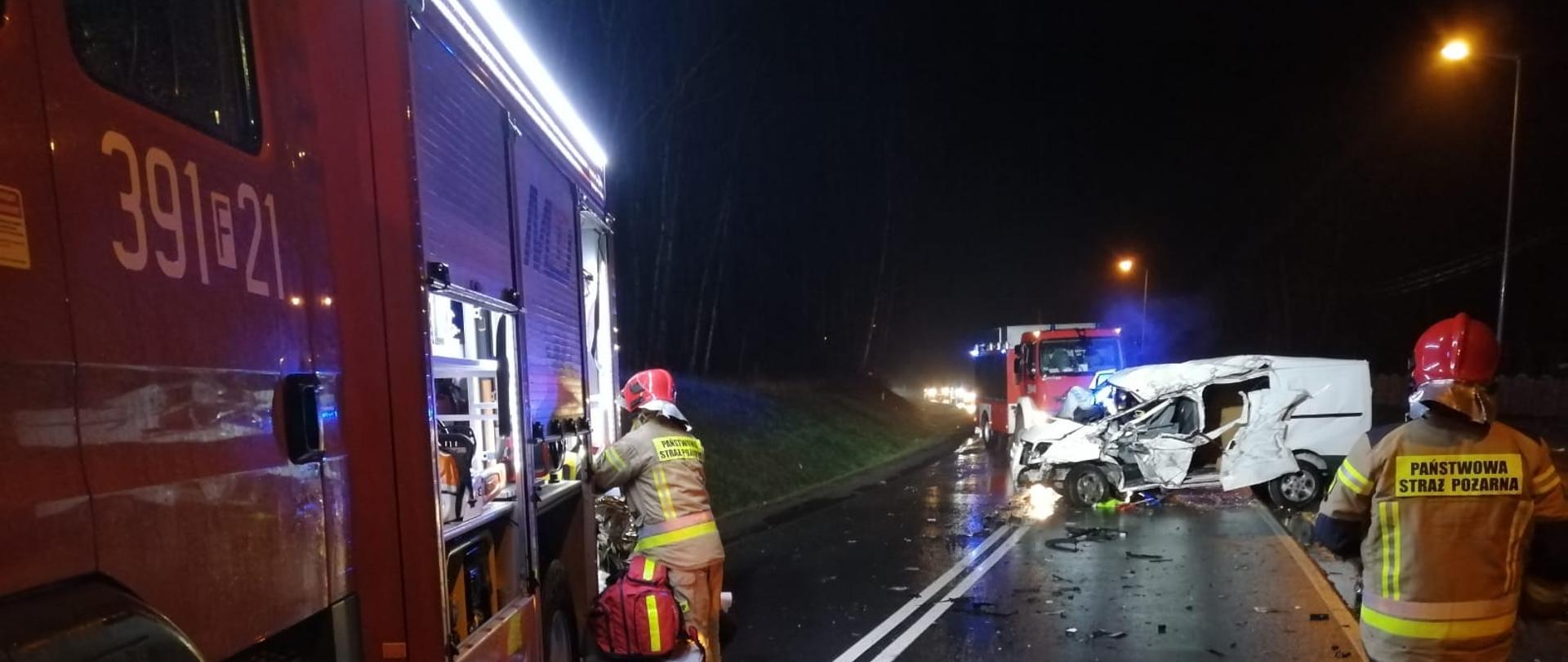 Strażacy PSP prowadzący działania na jezdni. Widoczne dwa samochody pożarnicze oraz rozbity, stojący na jezdni, biały samochód dostawczy.