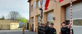 Na prawej stronie zdjęcia za niskim żywopłotem znajduję się biało – czerwony maszt na który jest wciągana Flaga Państwowa przez stojących 3 strażaków (Poczet flagowy) w ubraniach koszarowych. W taki sposób że środkowy strażak ciągnie za linke na której jest zawieszona flaga. A pozostali dwaj stoją po jego bokach i salutują. Flaga w chwili zdjęcia jest w połowie masztu. Na budynku obok widać zawieszone 3 flagi, figurkę św. Floriana i Duże Logo PSP. Pod flagami stoją plecami do budynku strażacy zmiany służbowej w dwuszeregu. Dowódca uroczystości stoi przed dwuszeregiem i salutuje. Najdalej na zdjęciu znajduję się obok budynku salutujący strażak obsługujący nagłośnienie (pojedyncza kolumna głośnikowa). 