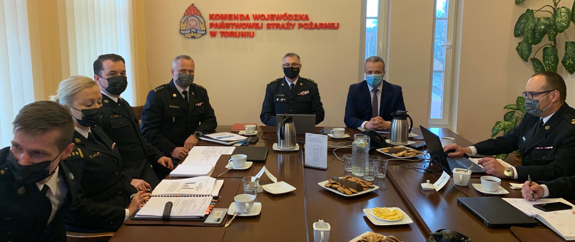 Zdjęcie przedstawia sześciu strażaków (w tym jedną kobietę) ubranych w mundury galowe siedzących wokół prostokątnego brązowego stołu. Oprócz nich w spotkaniu udział bierze mężczyzna w garniturze. Na stole przed poszczególnymi osobami znajdują się komputery przenośne notatniki, ciastka filiżanki i termosy z gorącymi napojami. W tle pastelowa ściana na której wisi godło Polski, logo Państwowej Straży Pożarnej i czerwony napis „Komenda Wojewódzka Państwowej Straży Pożarnej”. 
