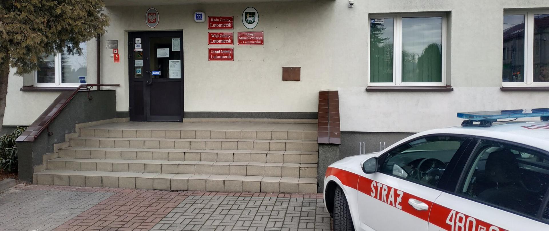 W tle wejście do budynku Urzędu Gminy w Lutomiersku, po prawej stronie kadru pojazd operacyjny Komendy Powiatowej Państwowej Straży Pożarnej w Pabianicach o numerze operacyjnym 480E90.