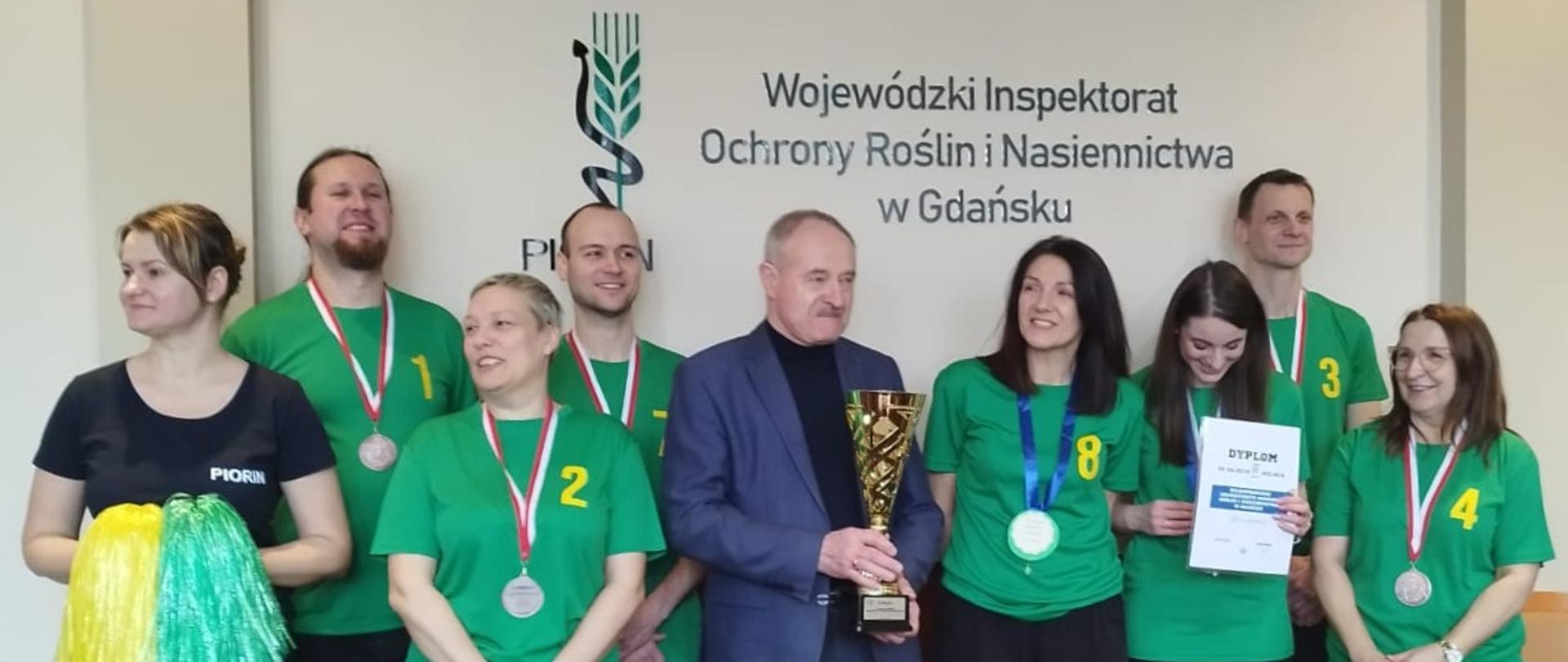 Grupa osób w zielonych koszulkach pozuje do zdjęcia z mężczyzną w garniturze trzymającym puchar w rękach 