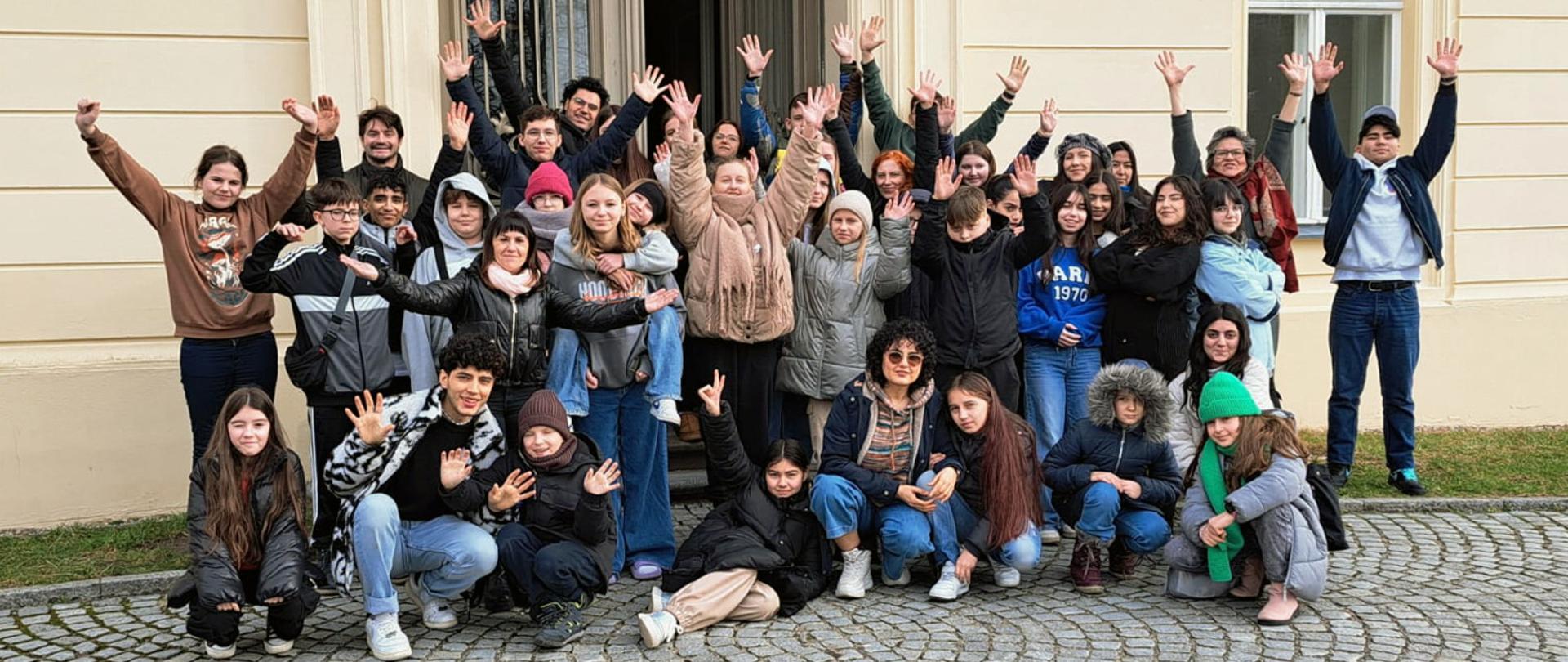 Na zdjęciu grupa kilkudziesięciu osób, dorosłych i dzieci. Osoby z przodu kucają i klęczą, osoby z tyłu stoją, część z osób ma uniesione ręce do góry. Za nimi jakiś budynek.