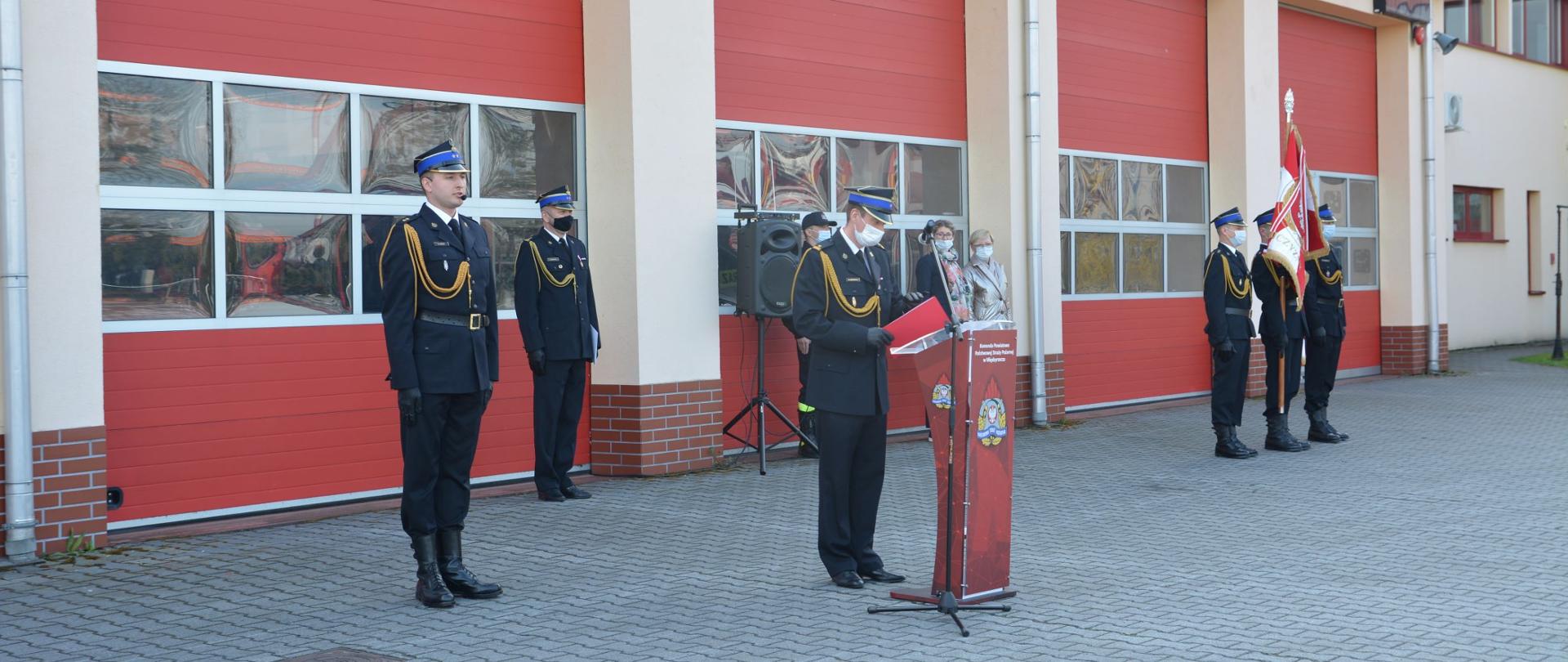 Zdjęcie przedstawia funkcjonariuszy PSP z Komendy Powiatowej w Międzyrzeczu oraz pracowników cywilnych podczas uroczystego apelu na placu przy remizie. Przemawia komendant powiatowy. Obok stoi poczet sztandarowy. W tle garaże dla pojazdów pożarniczych.