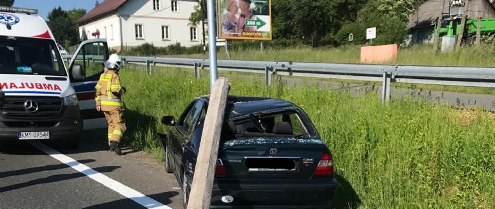 Zdjęcie przedstawia samochód osobowy w kolorze zielonym, który uderzył w betonowy słup oświetleniowy. Złamany słup leży na karoserii pojazdu. Z lewej strony stoi karetka pogotowia, a przed nią strażak w umundurowaniu specjalnym. W tle widoczne zabudowania miejscowości.