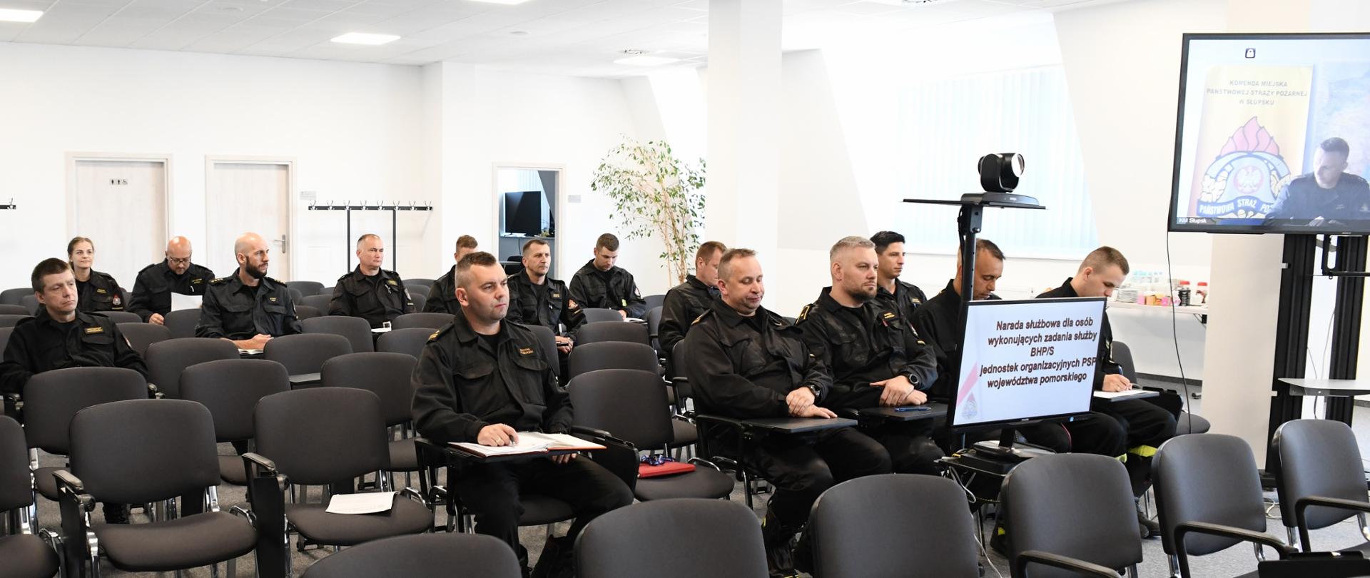 Funkcjonariusze Państwowej Straży Pożarnej w czarnych mundurach siedzą na krzesłach.