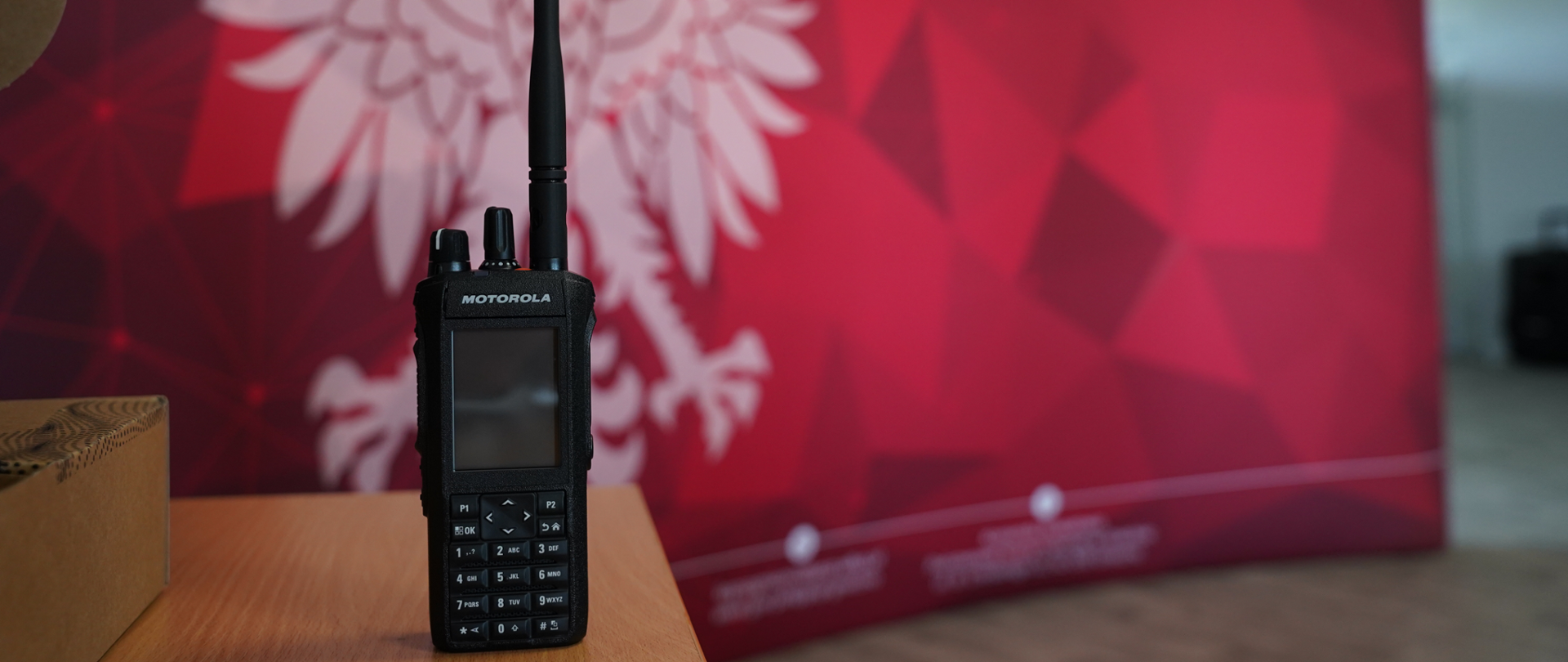 Na pierwszym planie na drewnianym stole stoi przenośne radio Motorola z długą anteną. W tle znajduje się czerwone tło z białym wizerunkiem orła oraz napisami Państwowej Straży Pożarnej.