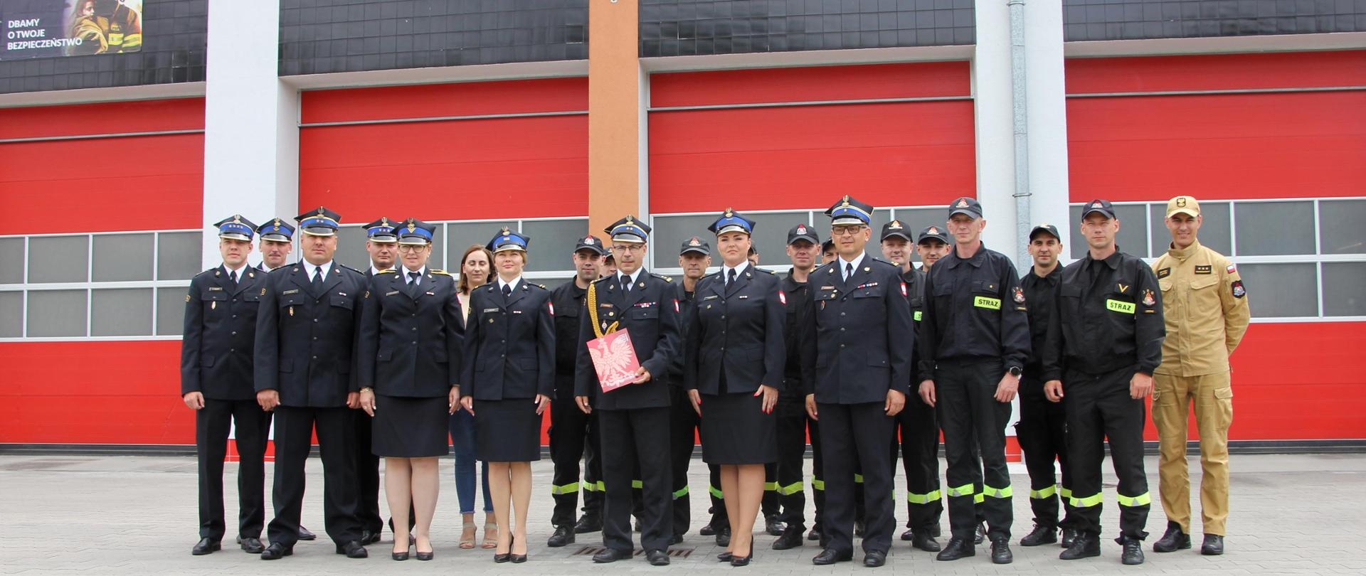 Strażacy w mundurach stoją w dwuszeregu. W tle widoczna komenda miejska.