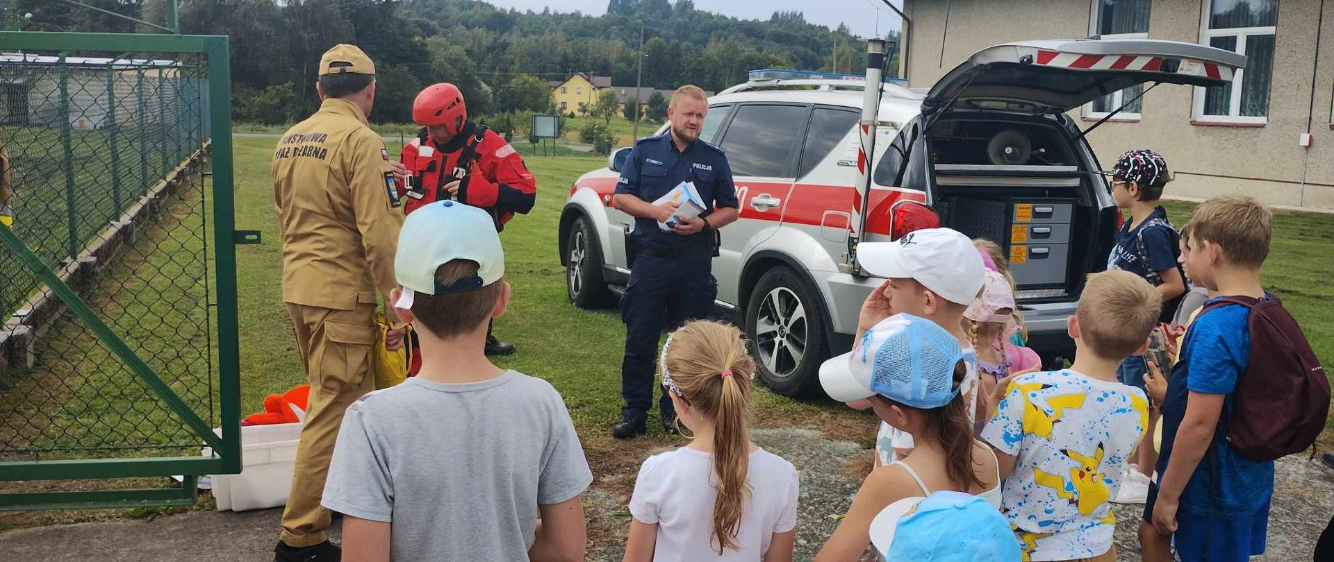Kolorowa fotografia wykonana w pogodny dzień na akwenie. Na pierwszym planie dzieci które słuchają strażaka ubranego w ubraniu służbowym. W tle policjant i samochód operacyjny straży pożarnej oraz strażak ochotnik ubrany w ubranie do pracy w wodzie.
