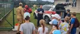 Kolorowa fotografia wykonana w pogodny dzień na akwenie. Na pierwszym planie dzieci które słuchają strażaka ubranego w ubraniu służbowym. W tle policjant i samochód operacyjny straży pożarnej oraz strażak ochotnik ubrany w ubranie do pracy w wodzie.