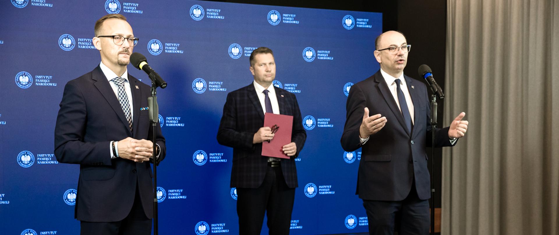 Minister Czarnek stoi za dwoma mężczyznami w garniturach, którzy mają przed sobą mikrofony na stojakach, trzyma w ręku fioletową teczkę, za nimi niebieska ściana.