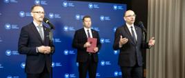 Minister Czarnek stoi za dwoma mężczyznami w garniturach, którzy mają przed sobą mikrofony na stojakach, trzyma w ręku fioletową teczkę, za nimi niebieska ściana.