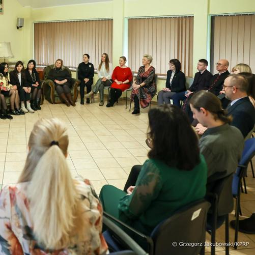 Około 20 osób siedzi w kółku na krzesłach patrząc na ekran, na którym uruchomiony jest program videokonferencyjny.