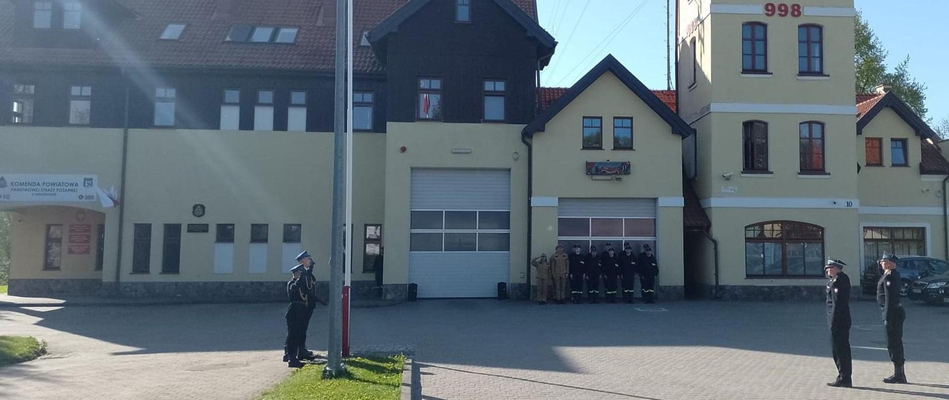 Strażacy stoją na placu podczas uroczystej zmiany służby i podnoszą flagę z okazji Dnia Flagi Rzeczypospolitej Polskiej