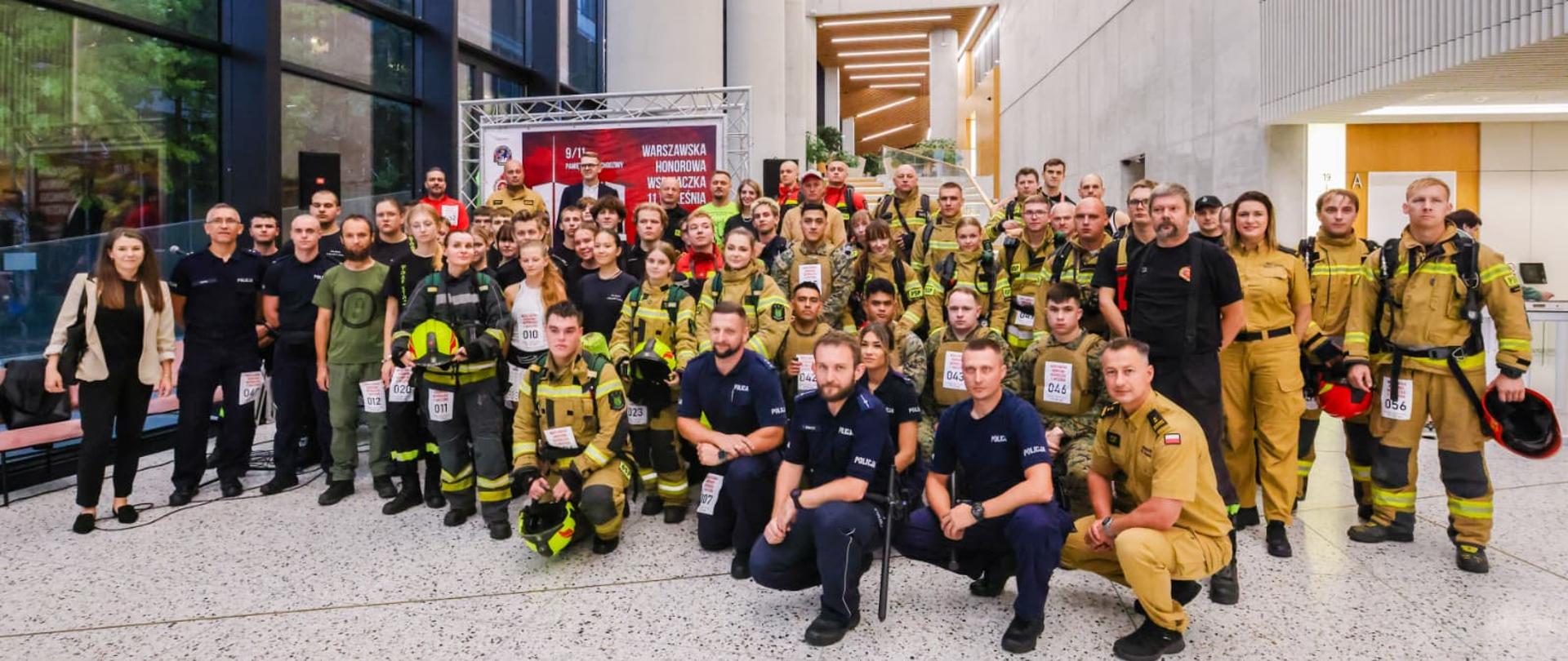 Grupa osób w mundurach strażackich i policyjnych, a także kilku cywilów, pozuje do zdjęcia w holu dużego, nowoczesnego budynku. Wszyscy są uśmiechnięci i wydają się zadowoleni, tuż przed wejściem po schodach.