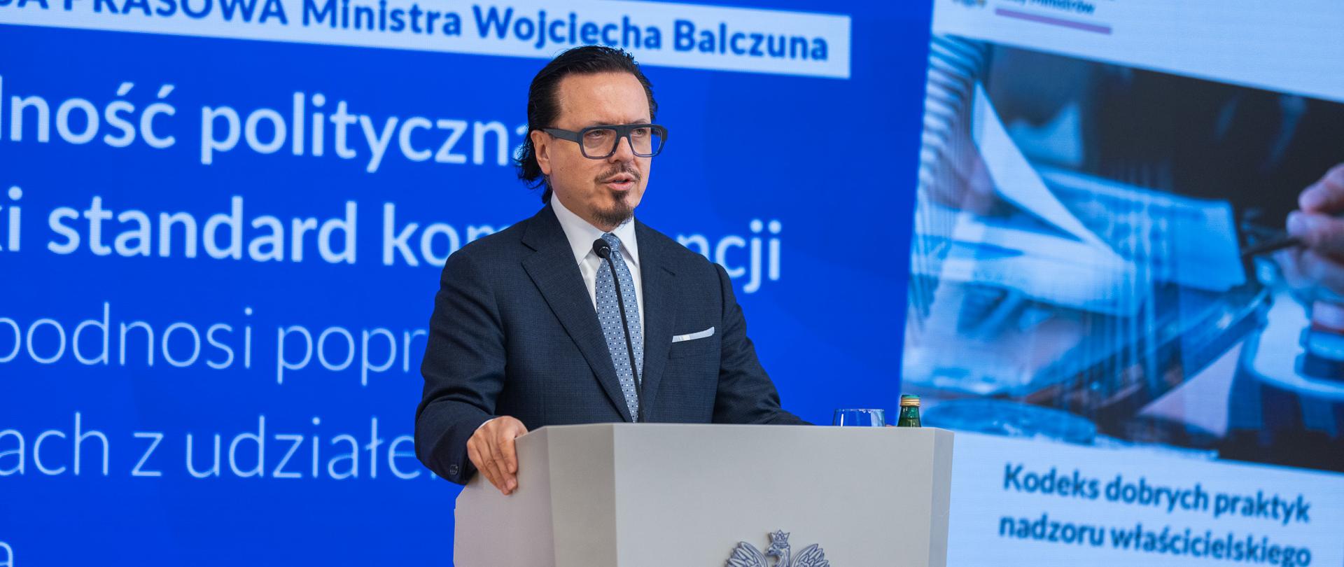 Minister Wojciech Balczun stoi za mównicą. W tle ekran. 