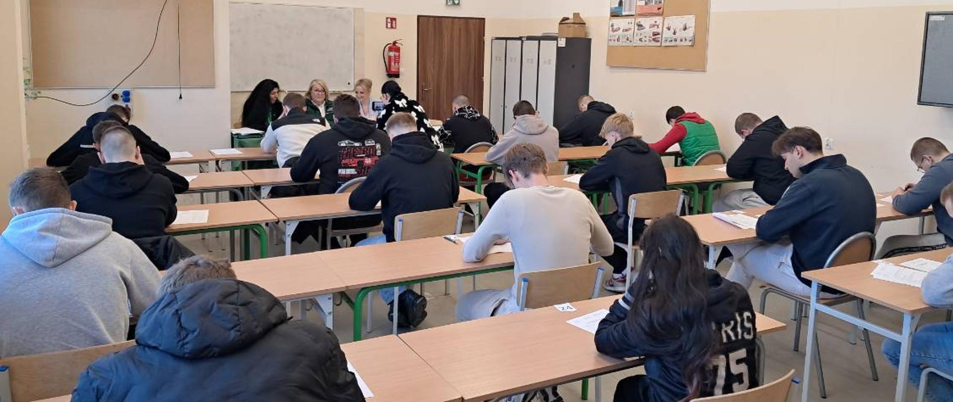 W klasie uczniowie siedzą w ławkach i piszą test pisemny. Z przodu sali, przy tablicy, siedzi komisja egzaminacyjna, która nadzoruje przebieg egzaminu.