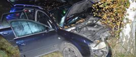 Wypadek w Stwolnie. Na pierwszym planie stojący w rowie czarny samochód osobowy, który czołowo uderzył w drzewo - widziany od strony prawego przedniego narożnika. Auto ma uszkodzony przód. Otwarta jest pokrywa silnika oraz drzwi i klapa bagażnika. 