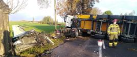 Na zdjęciu samochód osobowy znajdujący się poza obrysem jezdni po uderzeniu w drzewo. Na jezdni leży przewrócony samochód ciężarowy również po uderzeniu w drzewo.