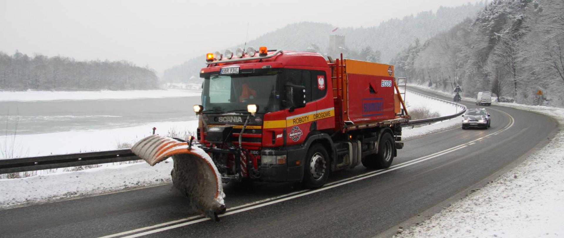 Zakręt ośnieżonej drogi, przebiegającej nad rzeką. Na pierwszym planie czerwona samochód ciężarowy z pługiem śnieżnym umocowanym z przodu i zasobnikiem na solankę umocowanym w części ładunkowej. W tle ośnieżone drzewa, baszta zamku z biało-czerwoną flagą