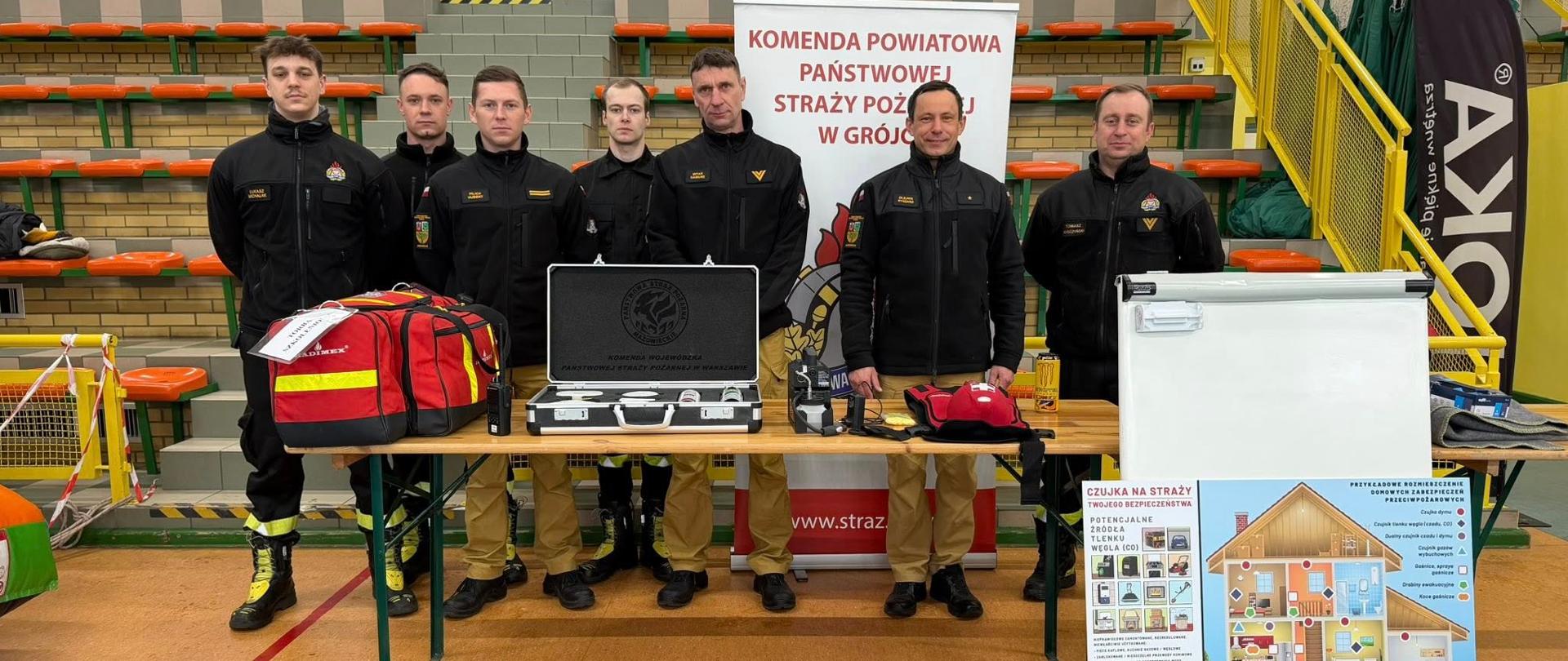 Na zdjęciu znajduje się grupa siedmiu osób stojących za dwoma stołami w hali sportowej. Wszyscy mają na sobie podobne, ciemne bluzy oraz spodnie robocze, co sugeruje, że są członkami jednej służby.
Na stołach leży sprzęt ratowniczy: torby medyczne, walizka z czujnikami, kaski, rękawice oraz inne elementy wyposażenia. Przed stołami, na podłodze, rozłożone są nosze ratownicze i dodatkowy sprzęt techniczny.
Za grupą stoi baner z napisem „Komenda Powiatowa Państwowej Straży Pożarnej w Grójcu” oraz materiały edukacyjne dotyczące ochrony przeciwpożarowej.
