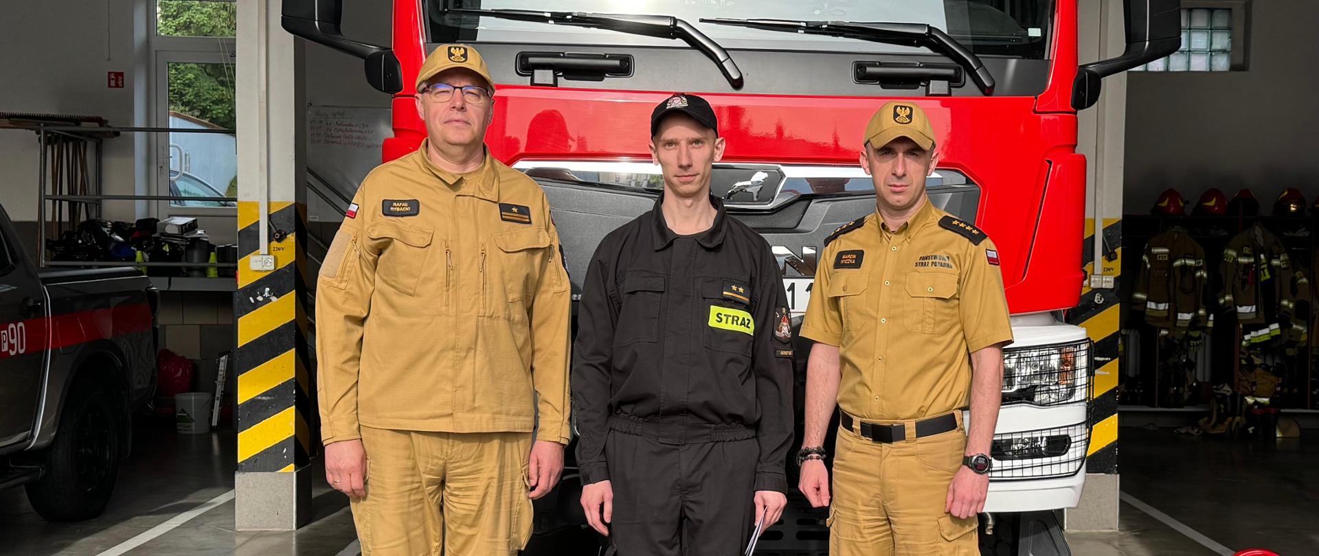 Komendanta, dowódcę jednostki ratowniczo-gaśniczej oraz wyróżnionego strażaka stojących obok siebie i pozujących do zdjęcia. Stoją oni przed wozem strażackim.