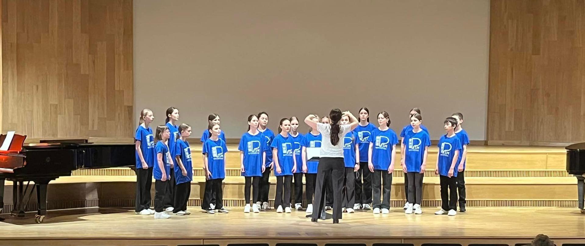Grupa uczniów w niebieskich koszulkach z logo szkoły muzycznej stojących na scenie sali koncertowej na tle biało-beżowej ścianie.