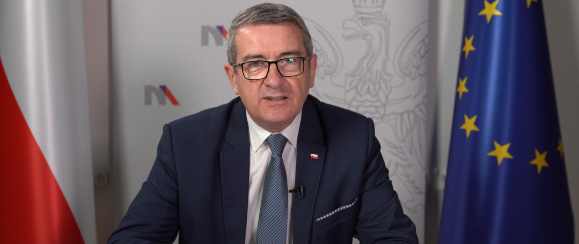 Minister Wojciech Murdzek siedzi za stołem, za nim flagi Polski i UE.