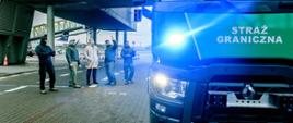 Na pierwszym planie zdjęcia, po jego prawej stronie znajduje się nowoczesny ambulans pirotechniczny marki Renault, przekazany funkcjonariuszom z Grupy Interwencji Specjalnych Placówki Straży Granicznej Warszawa-Okęcie. Wóz oklejony jest zieloną folią z białym napisem "Straż Graniczna" i w trakcie wykonania zdjęcia "wysyła" sygnały świetlne jako pojazd uprzywilejowany. W tle, po lewej stronie zdjęcia stoi pięciu mężczyzn: jeden ubrany w beżowy płaszcz i czterech w mundury. Jeden z mężczyzn ubranych w mundur gestykuluje, wskazując ręką w kierunku wozu. 