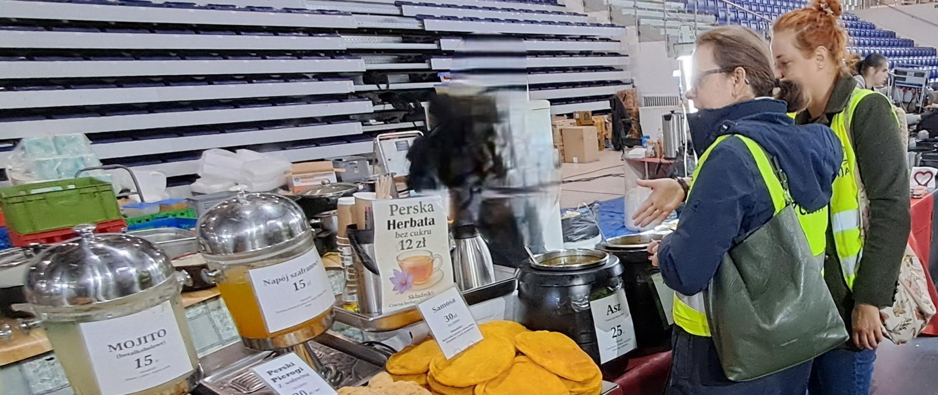 Dwie inspektorki IJHARS stoją przy stoisku z żywnością azjatycką, oglądając produkty. W tle widoczne są inne stoiska targowe z różnorodną żywnością.