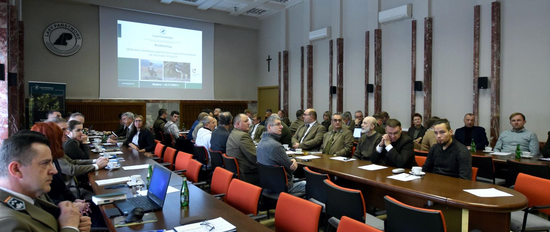 Sala konferencyjna, prezentacja, przy długich stołach siedzą uczestnicy konferencji