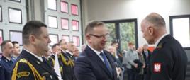 Prezydent Miasta gratuluje Komendantowi ściskając jego dłoń, mówi do niego, patrzą na siebie. Po jego lewej stronie stoi Komendant Wojewódzki, a w tle za nimi inni strażacy i policjanci w mundurach oraz osoby cywilne.
