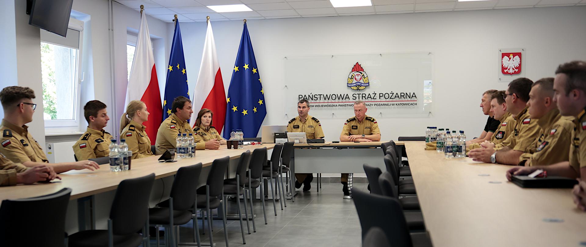 Zdjęcie przedstawia przestronną, jasną salę konferencyjną w Komendzie Wojewódzkiej Państwowej Straży Pożarnej w Katowicach. Przy dużym, ustawionym w kształcie litery „U” stole siedzi kilkunastu funkcjonariuszy Państwowej Straży Pożarnej w umundurowaniu służbowym. Uczestnicy spotkania zwróceni są w stronę dwóch strażaków siedzących przy centralnym stole, wśród nich jest śląski komendant wojewódzki starszy brygadier Damian Legierski. Na środku sali, za prowadzącymi, znajduje się laptop oraz sprzęt konferencyjny. Na ścianie widoczne jest logo Państwowej Straży Pożarnej i napis „Państwowa Straż Pożarna – Komenda Wojewódzka Państwowej Straży Pożarnej w Katowicach”. Po prawej stronie umieszczone jest godło Polski. W tle znajdują się cztery stojące flagi – dwie biało-czerwone oraz dwie flagi Unii Europejskiej. Wokół stołu ustawione są czarne krzesła, a na blatach znajdują się butelki z wodą, szklanki, dokumenty i urządzenia elektroniczne. 