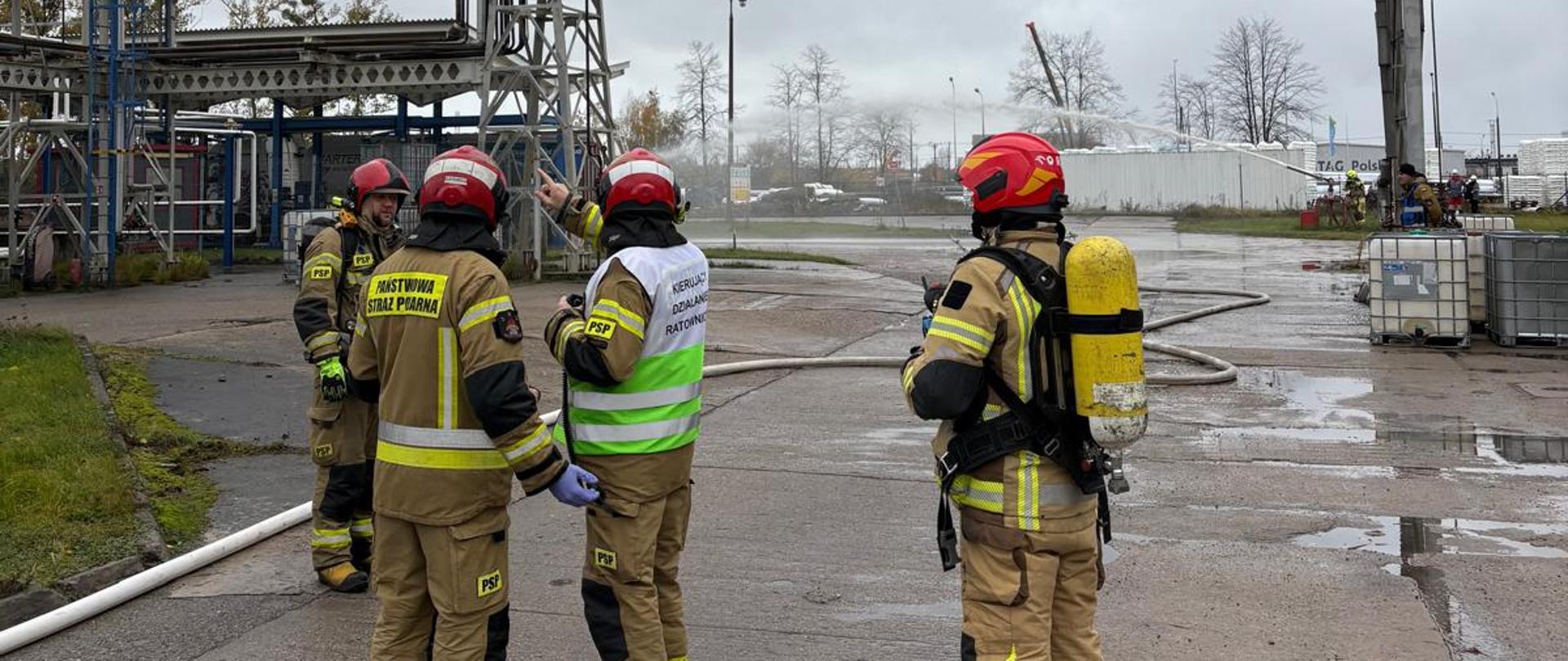 Na zdjęciu czterech strażaków PSP w ubraniach specjalnych. Jeden z nich w biało-zielonej kamizelce z napisem „KIERUJĄCY DZIAŁANIEM RATOWNICZYM” wskazuje na metalową konstrukcję znajdującą się za strażakami w górnej części zdjęcia. Ponad nimi pochmurne niebo. 