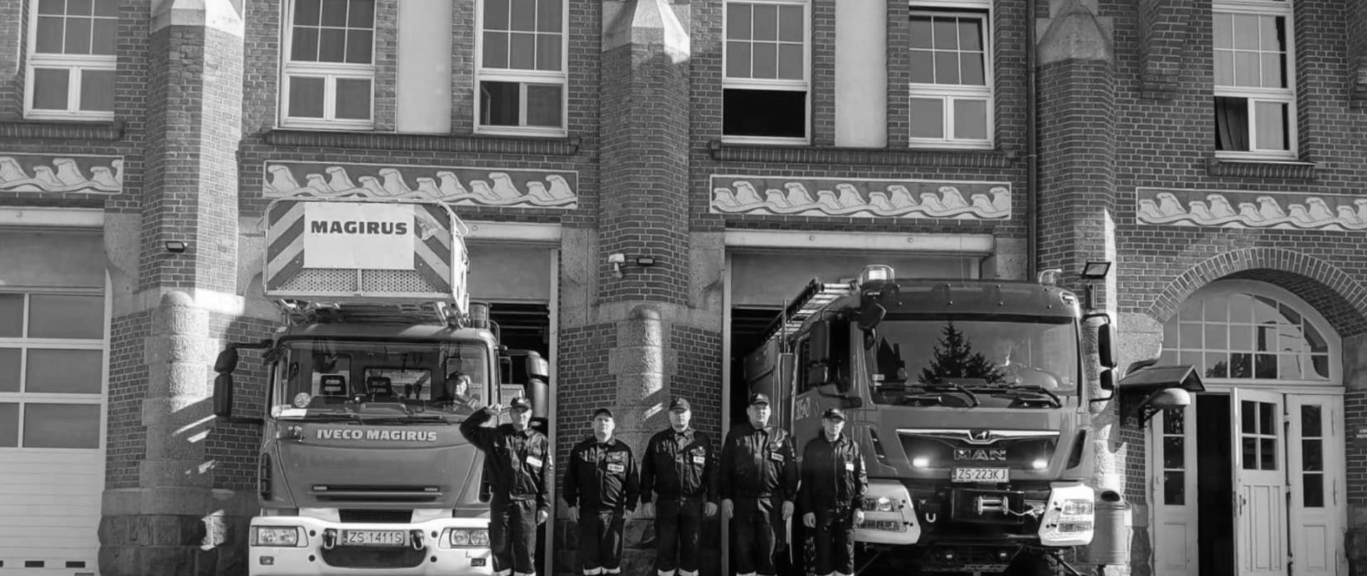Obrazek przedstawia sześciu strażaków ustawionych w szeregu przed budynkiem straży pożarnej, pomiędzy dwoma zaparkowanymi wjazdami garażowymi. Po lewej stronie stoi czerwony wóz strażacki Iveco Magirus, a po prawej nowszy wóz marki MAN. Budynek jest z cegły, z oknami na piętrze, a podłoga wykonana jest z kostki brukowej. Strażacy mają na sobie mundury i stoją na baczność, jeden z nich salutuje.