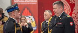 Dwóch mężczyzn w mundurach ściska sobie dłonie podczas oficjalnej uroczystości. W tle widać czerwone dekoracje i kilka osób obserwujących wydarzenie.