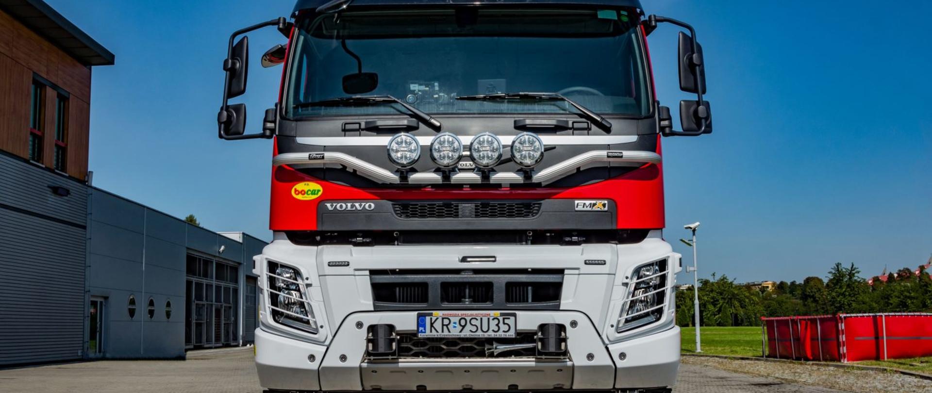 Duży czerwony pojazd gaśniczy na tle niebieskiego nieba. Obok widoczny budynek komendy straży oraz zbiornik wodny. Pojazd jest przedstawiony od frontu, widać tablicę rejestracyjną . 