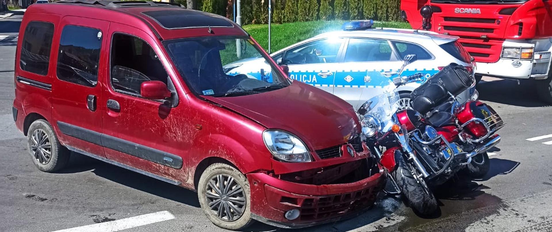 Na zdjęciu widać samochód i motocykl biorący udział w wypadku na skrzyżowaniu. W tle widać samochód policyjny i strażacki oraz bloki.