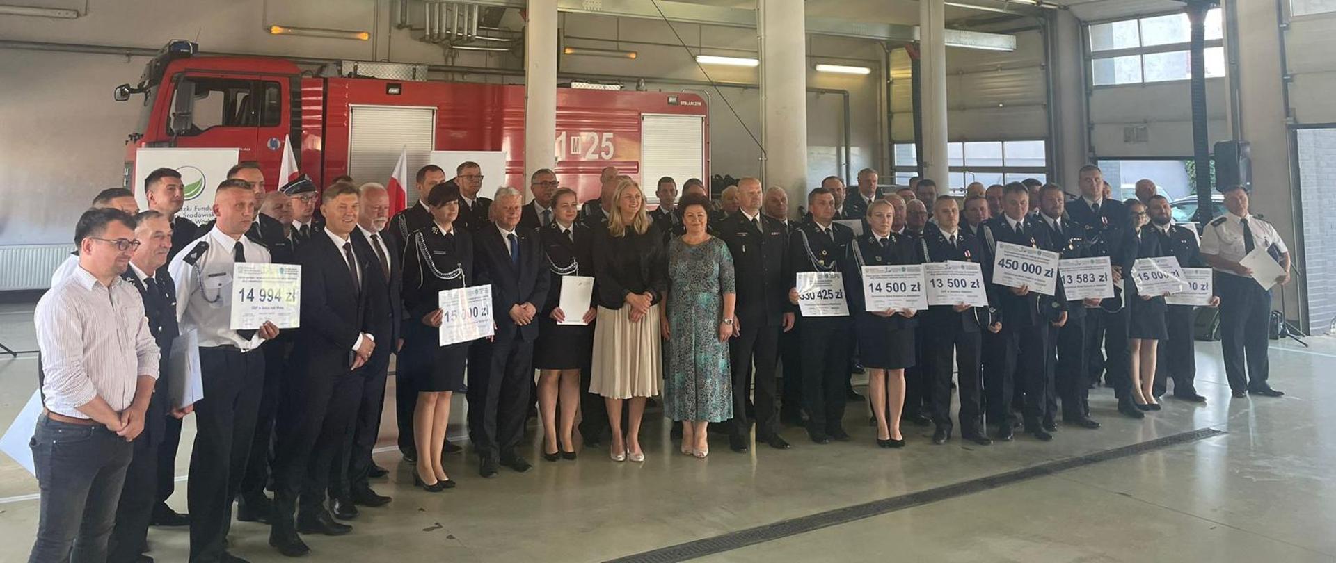 Zdjęcie wykonane na terenie obiektu jednostki ratowniczo – gaśniczej Państwowej Straży Pożarnej numer 1 w Radomiu. Na zdjęciu widać odbywającą się uroczystość wręczenia aktów dofinansowania w formie promes dla ochotniczych straży pożarnych. Dofinansowanie pochodzi z wojewódzkiego funduszu ochrony środowiska i gospodarki wodnej w warszawie. Na pamiątkowym zdjęciu wykonanym w garażu jednostki widać przybyłych na uroczystość gości posłów, parlamentarzystów, funkcjonariuszy Państwowej Straży Pożarnej oraz członków ochotniczych straży pożarnych. Druhowie w rękach trzymają pamiątkowe promesy na których wypisane są kwoty przyznanych dofinansowań. W tle ustawiony jest pojazd pożarniczy w kolorze czerwonym. 