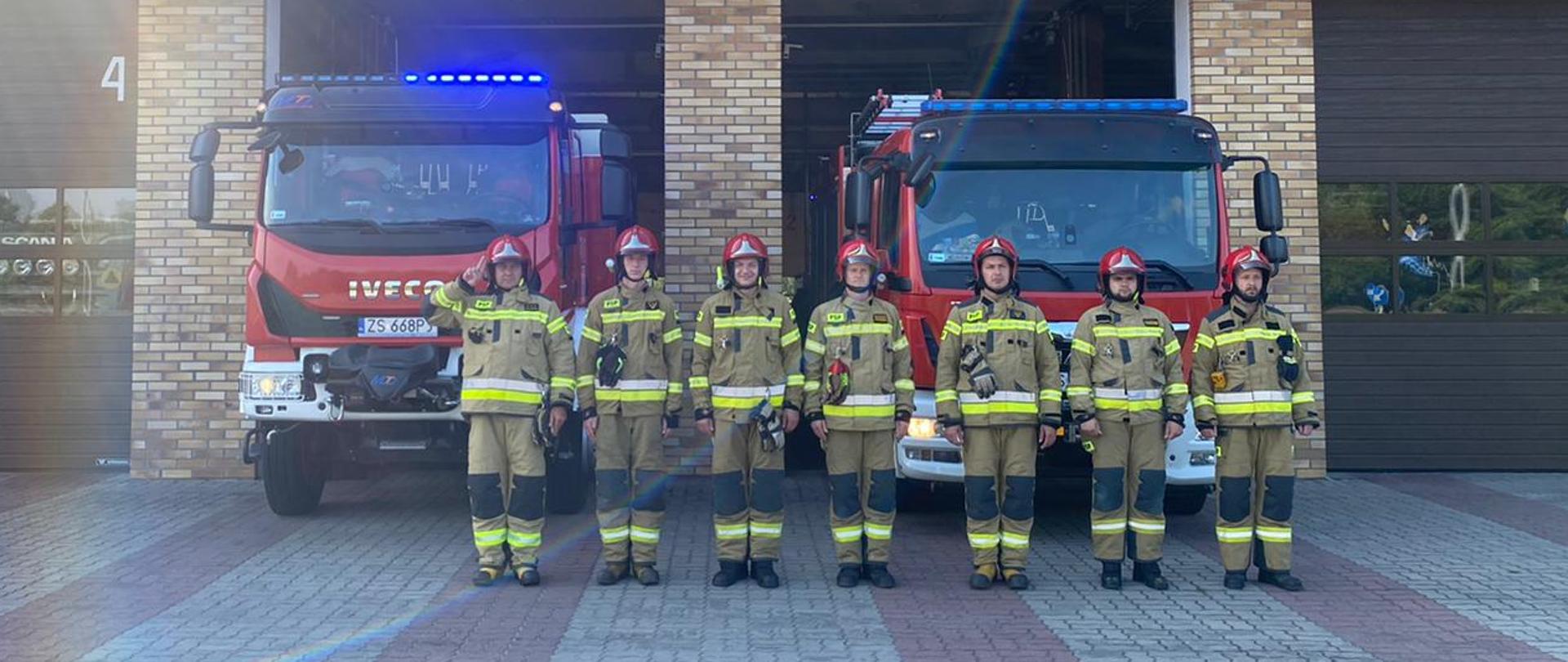 Strażacy stojący w rzędzie, za nimi samochody pożarnicze z włączonymi światłami