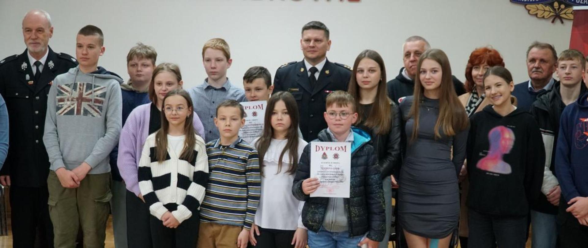 Zdjęcie przedstawia Komendanta Powiatowego Państwowej Straży Pożarnej w Zambrowie brygadiera Andrzeja Kalinowskiego ubranego w mundur wyjściowy oraz Prezesa Zarządu Oddziału Powiatowego Związku Ochotniczych Straży Pożarnych Rzeczpospolitej Polskiej w Zambrowie druha Marka Adama Komorowskiego ubranego w mundur wyjściowy Ochotniczych Straży Pożarnych podczas pozowania do zdjęcia z uczestnikami etapu powiatowego do Ogólnopolskiego Turnieju Widzy Pożarniczej. Zdjęcie wykonano w jednym z pomieszczeń Komendy Powiatowej Państwowej Straży Pożarnej w Zambrowie w słoneczny dzień.