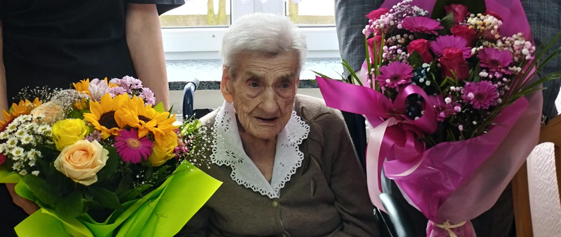 Na wózku siedzi starsza kobieta Jubilatka Pani Regina Kęsicka. Obok po prawej stronie stoi mężczyzna, który trzyma w rękach bukiet różowych kwiatów, a po lewej stronie stoi kobieta, która również trzyma w rękach bukiet kolorowych kwiatów. Wszyscy są przejęci i wzruszeni.