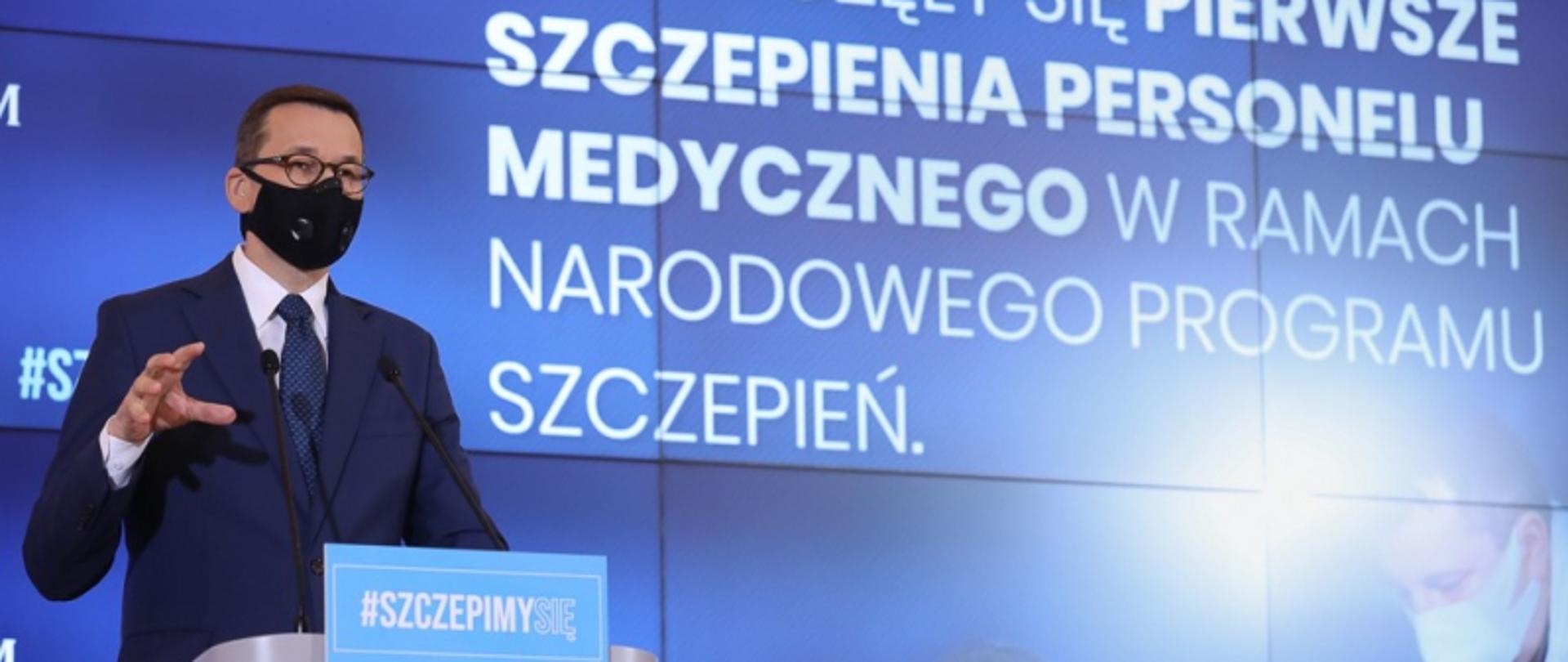Premier Morawiecki stoi przy mównicy za nim wyświetlany jest napis "szczepienia personelu medycznego w ramach narodowego programu szczepień"