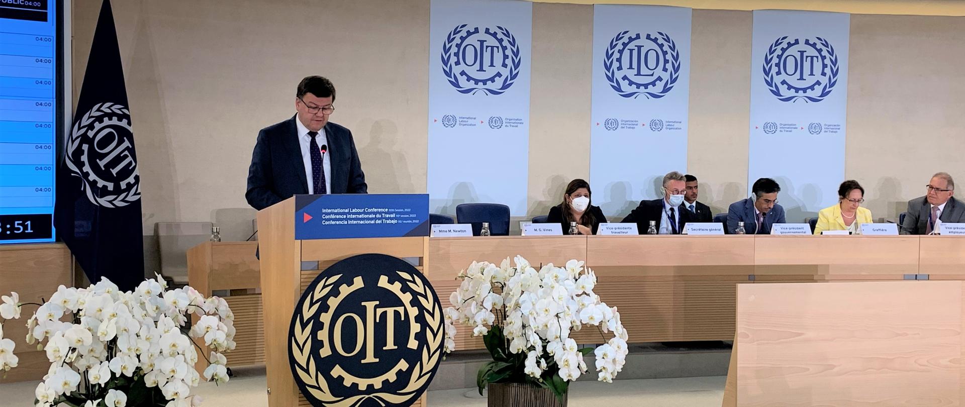Ambasador Zbigniew Czech przy mównicy z napisem OIT, przemawiający na sesji plenarnej Międzynarodowej Konferencji Pracy. Na prawo od Ambasadora siedzą uczestnicy Międzynarodowej Konferencji Pracy, za nimi zamieszczono na ścianie trzy duże bannery z napisem IOT. Po lewej i prawej stronie mównicy stoją okazałe storczyki, po lewej stronie stoi flaga Międzynarodowej Organizacji Pracy z napisem OIT.