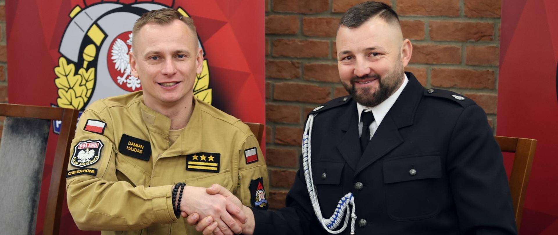 Siedzący za stołem konferencyjnym Zastępca Komendanta Centralnej Szkoły PSP w uścisku dłoni z druhem OSP, przed nimi leżace na blacie trzy smartfony, w tle po lewej baner z logo PSP