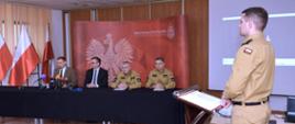 Na zdjęciu widzimy 4 mężczyzn podczas konferencji prasowej na tle czerwonej ścianki medialnej z napisem Państwowa Straż Pożarna i orłem w koronie. Mężczyźni siedzą za stołem i przed nimi mikrofony mediów.