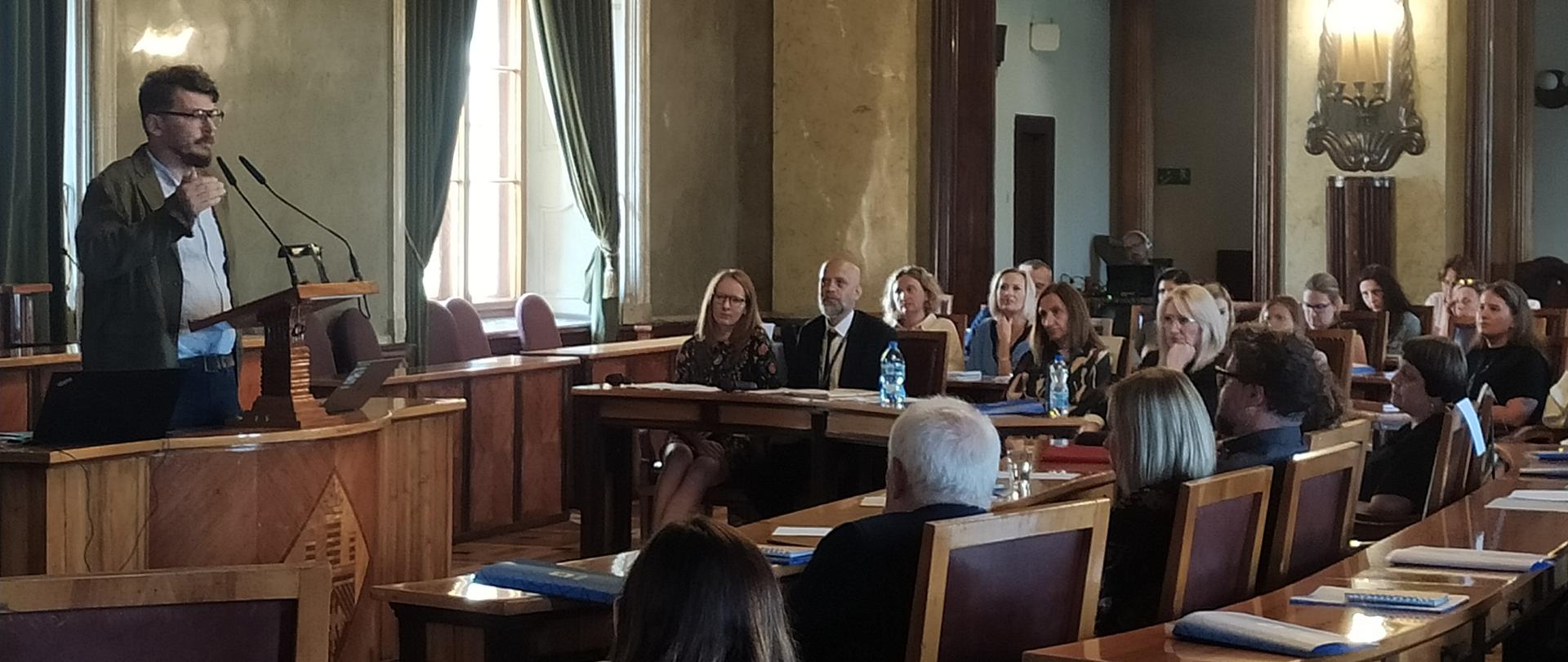 Na zdjęciu widać tłum ludzi uczestniczących w konferencji. Osoby siedzą przy długich stołach. Jedna osoba przemawia.