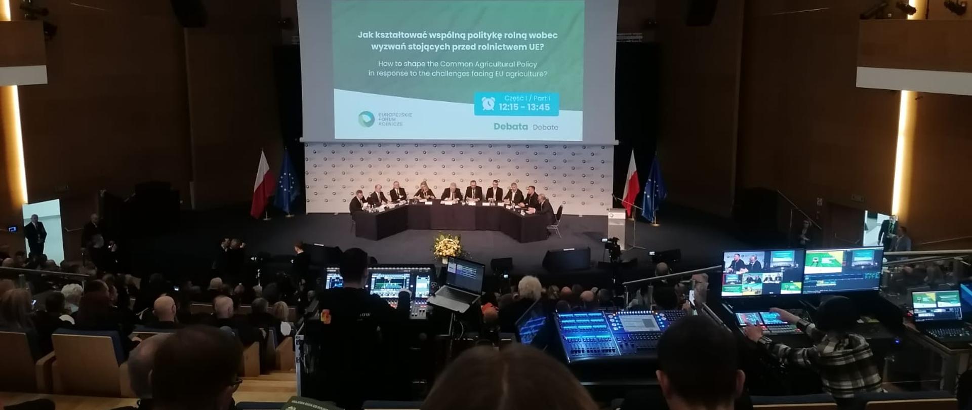Uczestnicy siedzący na sali konferencyjnej wraz z osobami prowadzącymi konferencję.