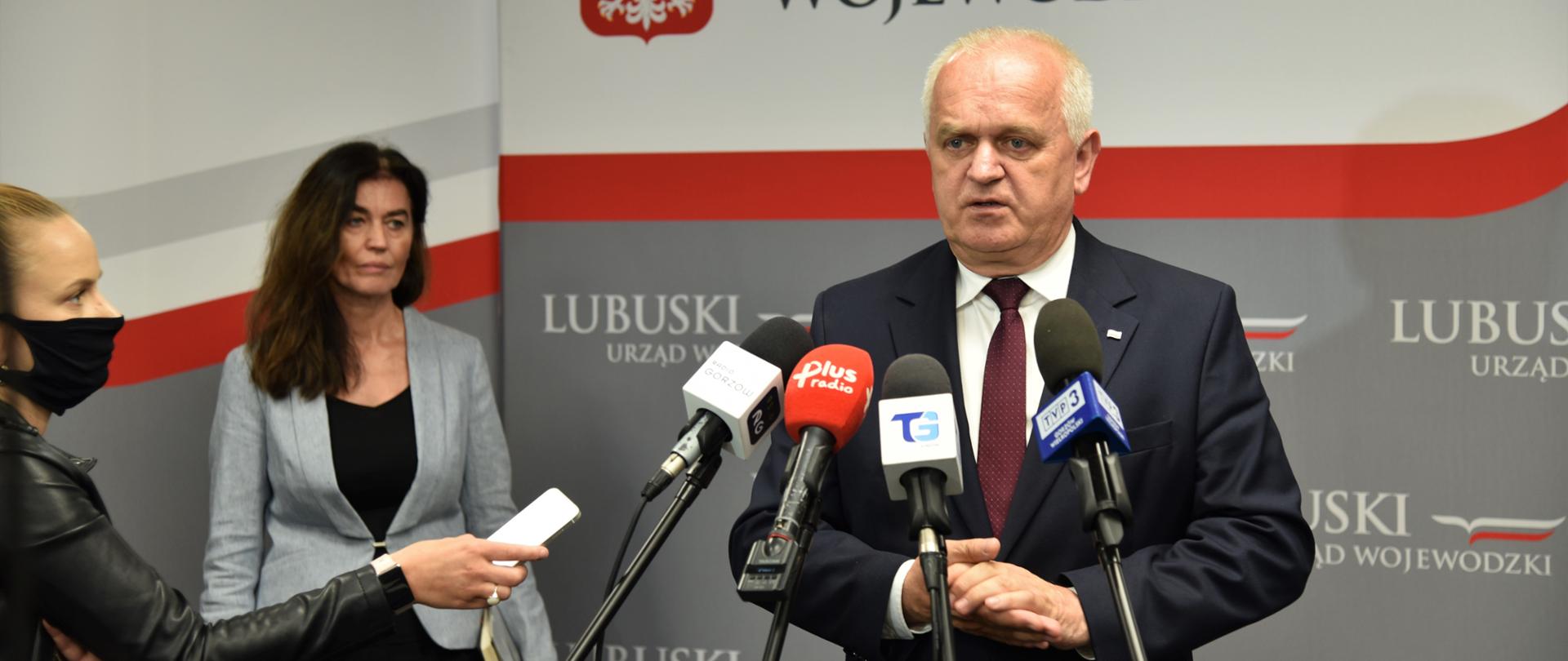 Wojewoda przemawia do mikrofonów ustawionych na statywach. W tle ścianki promocyjne urzędu wojewódzkiego. Po lewej stronie, za wojewodą stoi dyrektor polityki społecznej Grazyna Jelska