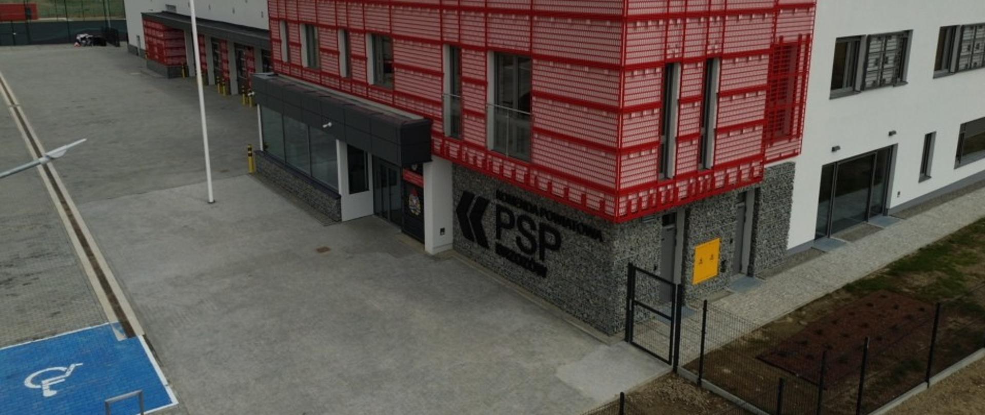 Budynek nowo wybudowanej Komendy Powiatowej PSP w Brzozowie. Zdjęcie wykonane z drona przedstawia plac na którym wydzielone jest miejsce parkingowe dla osób niepełnosprawnych. Budynek koloru białego z ażurowymi wykończeniami, przy wejściu napis wskazujący instytucję Komendy Powiatowej PSP w Brzozowie